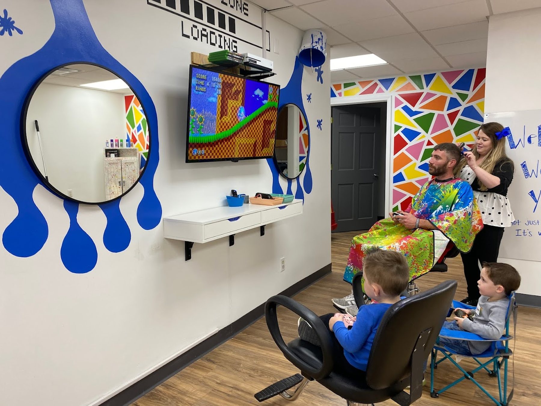 Two boys playing on facing video game stations under a wall painted with big blue paint drips, while a man gets his hair cut nearby.