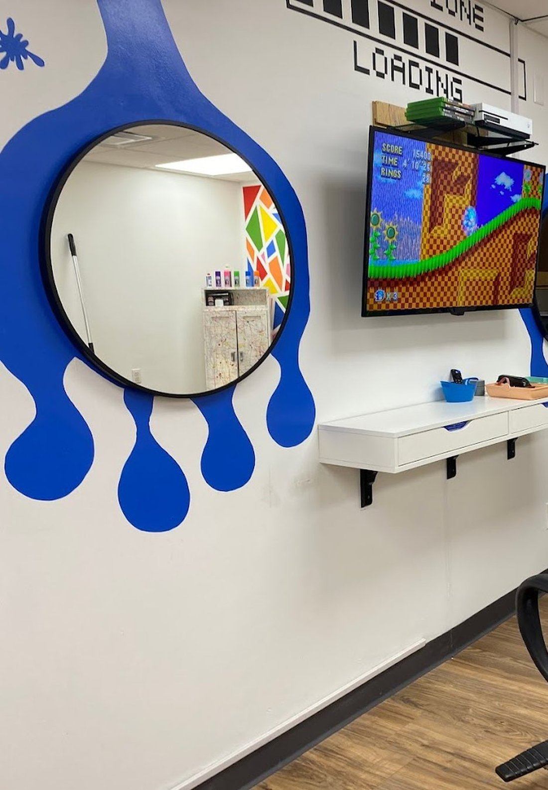 A round mirror mounted inside a large blue paint drip painted straight onto the salon wall, with a game console and screen on the shelf beside it.