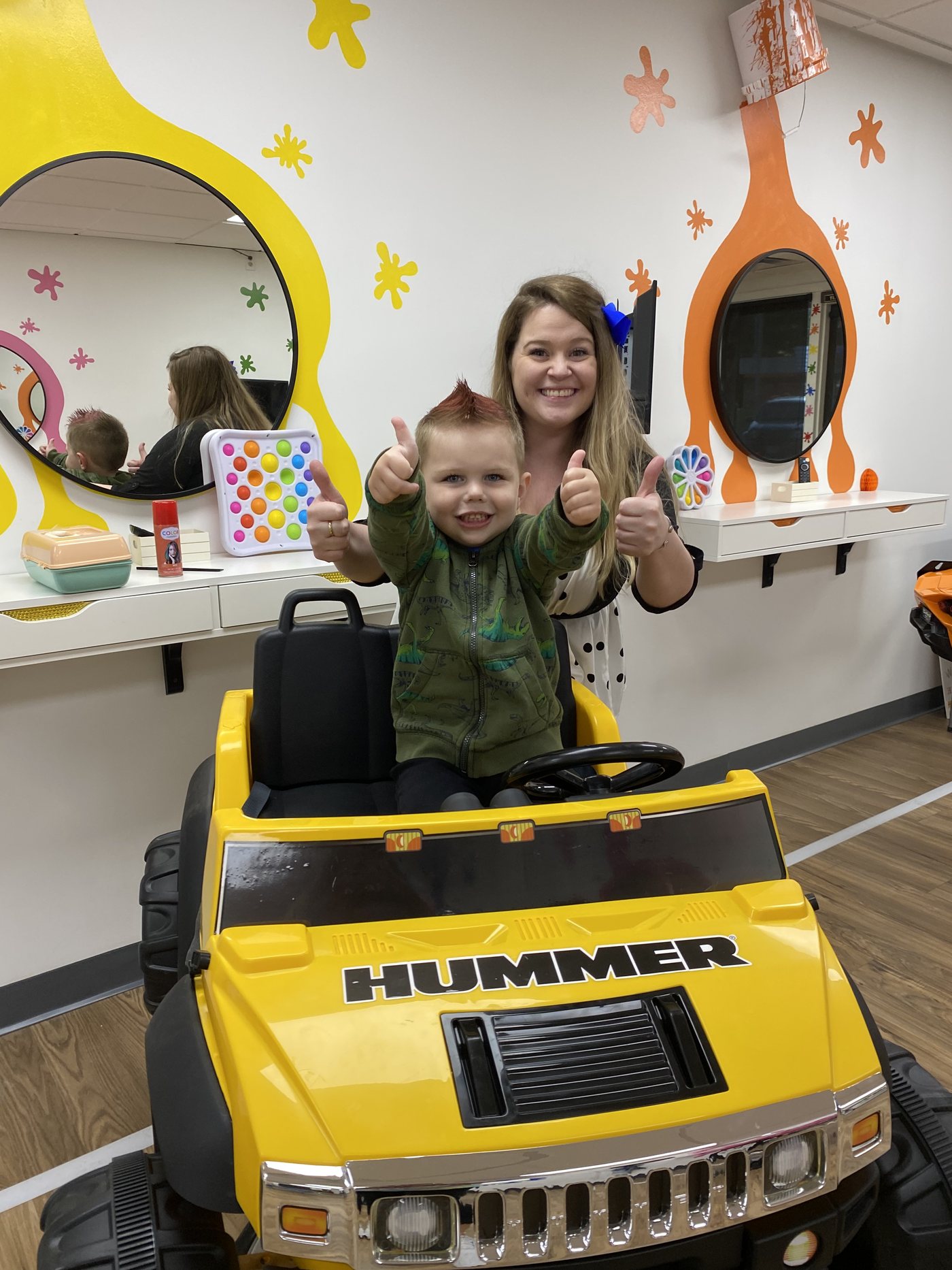A yellow ride-on truck parked at a styling station, a pink ride-on car behind it, and round mirrors cut into orange and yellow paint splats on the wall.