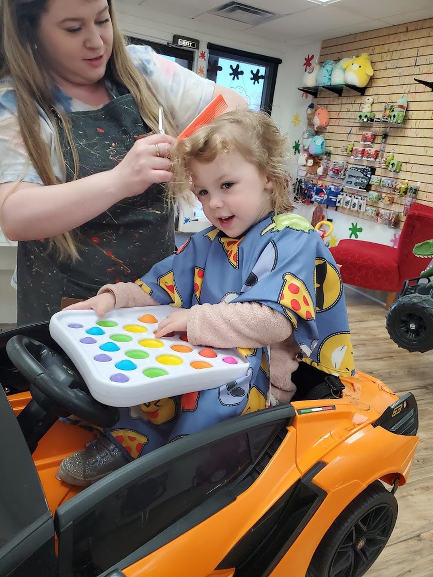 A toddler in an orange ride-on sports car holding a pop-it tray while a stylist trims her hair.