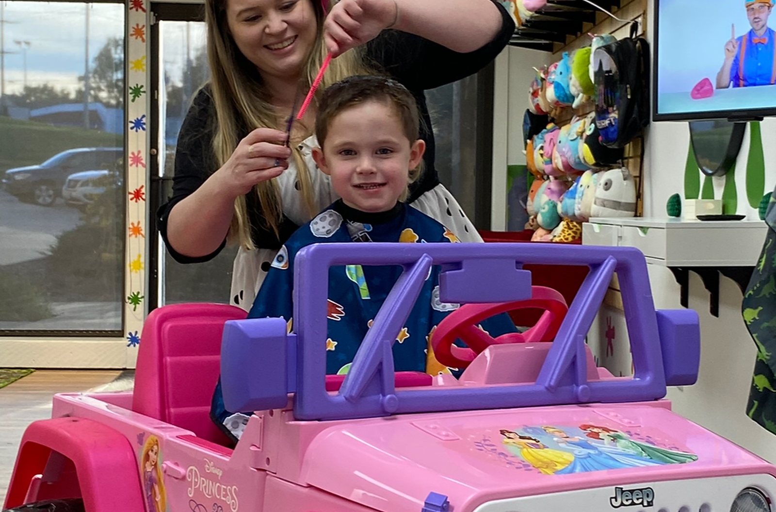 A child sitting in a pink ride-on car at the front window while a stylist combs his hair.