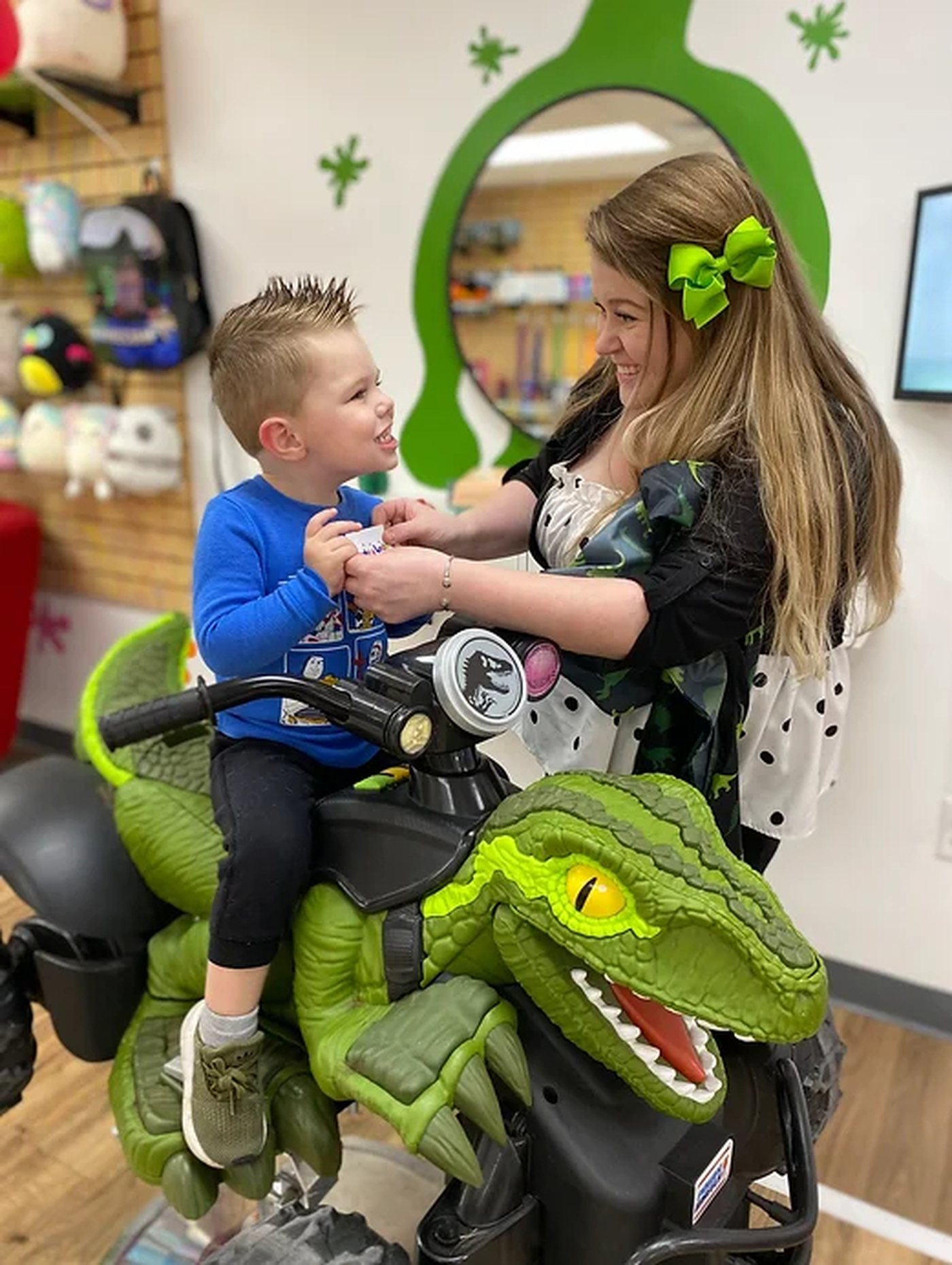 A boy sitting on a large green ride-on dinosaur, laughing with his stylist, against a green painted splat wall.